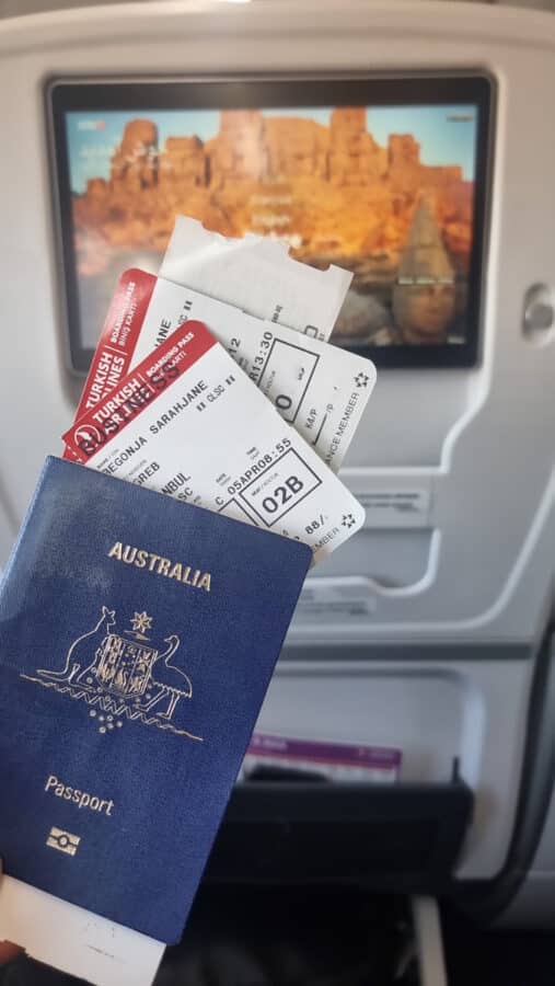 Australian passport and boarding passes in hand with an airplane seatback screen showing a scenic canyon in Turkey in the background.