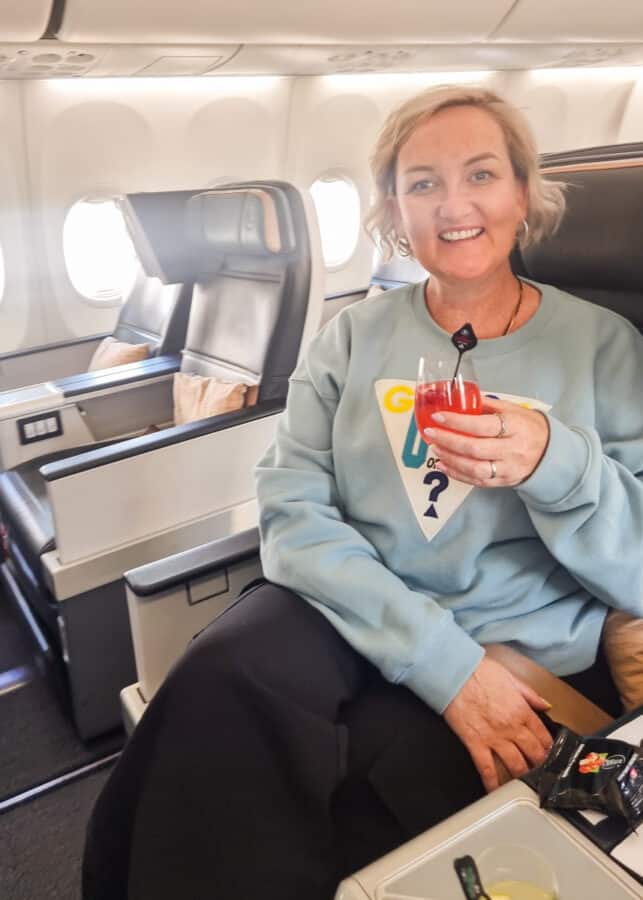 SJ with short blonde hair, smiling and holding a red drink, seated in a spacious airplane cabin en route to Turkey.