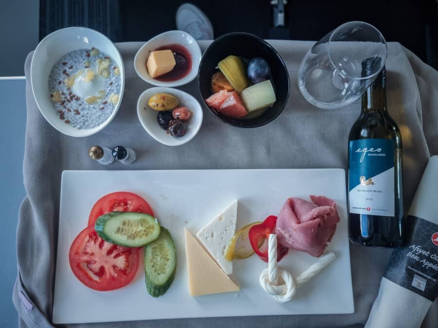 An airplane meal featuring a cheese plate, sliced cured meats, fresh vegetables, mixed fruit, yogurt with granola, and a bottle of red wine on a tray during a Turkey transport flight.