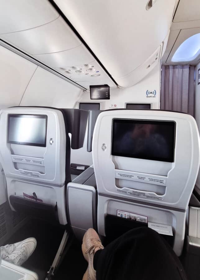 Interior of an airplane in Turkey showing two rows of seats with in-flight entertainment screens, featuring a view of the cabin aisle and overhead compartments.