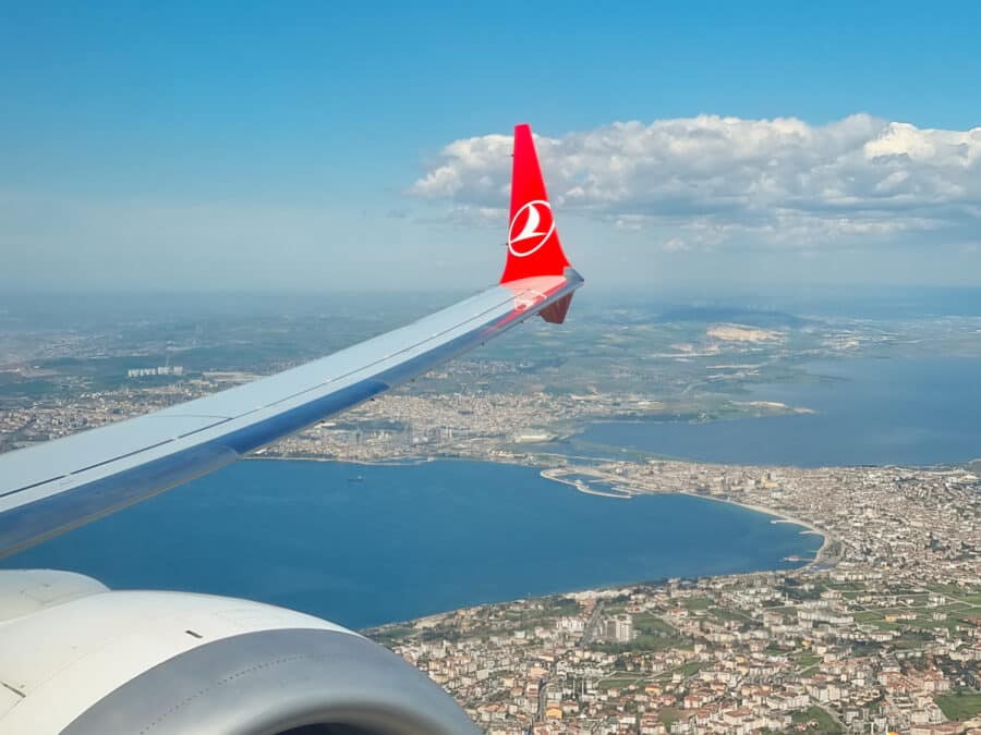 Aerial view from an airplane window showing a wing with a red logo, overlooking a coastal city in Turkey and blue sea.