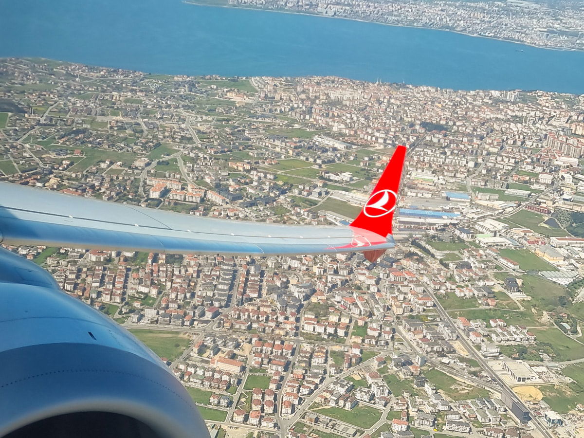 Aerial view from an airplane window showing a cityscape, coast, and a wing with a red Turkey Transport logo.