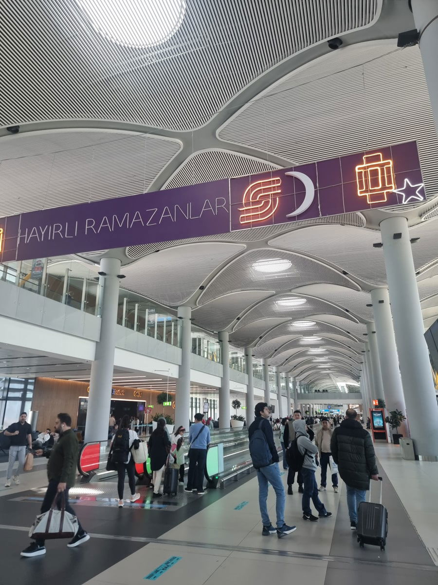 Interior of a busy airport terminal in Turkey, featuring travelers with luggage and a "hayırlı ramazanlar" sign overhead, meaning "happy Ramadan" in Turkish.