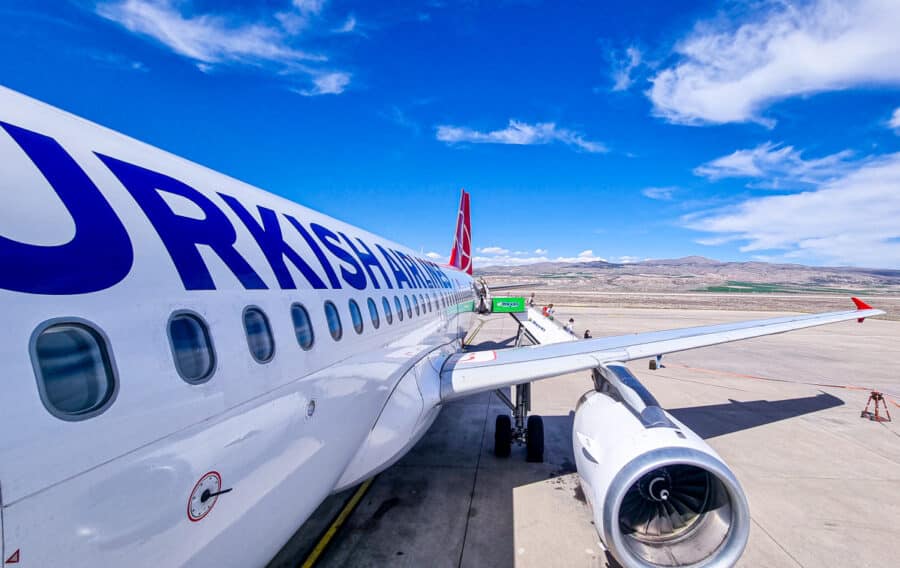 Flight ticket types - Passengers board a Turkey Transport plane on a sunny day with a clear blue sky and distant mountains in the background.