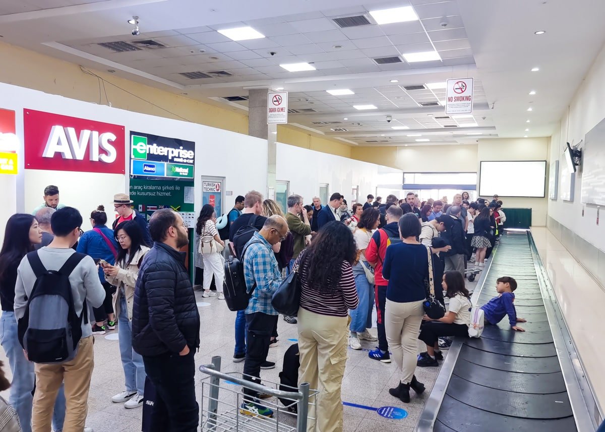 Crowded Nevşehir Kapadokya Airport baggage claim area in Turkey with people waiting for luggage, surrounded by car rental and transport advertisements.