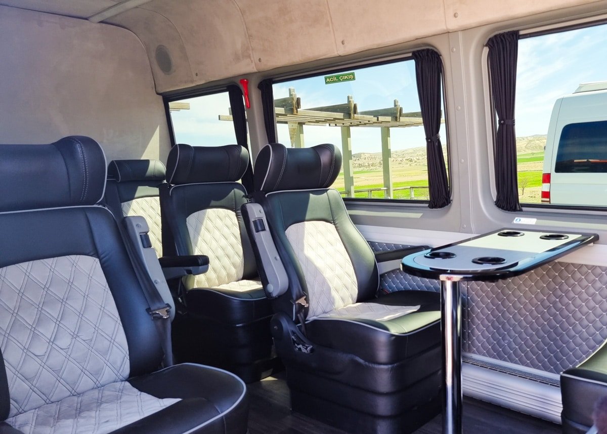 Interior of a luxury transport van with plush seating and a table, a scenic view of Turkey visible through the window, parked next to another vehicle.