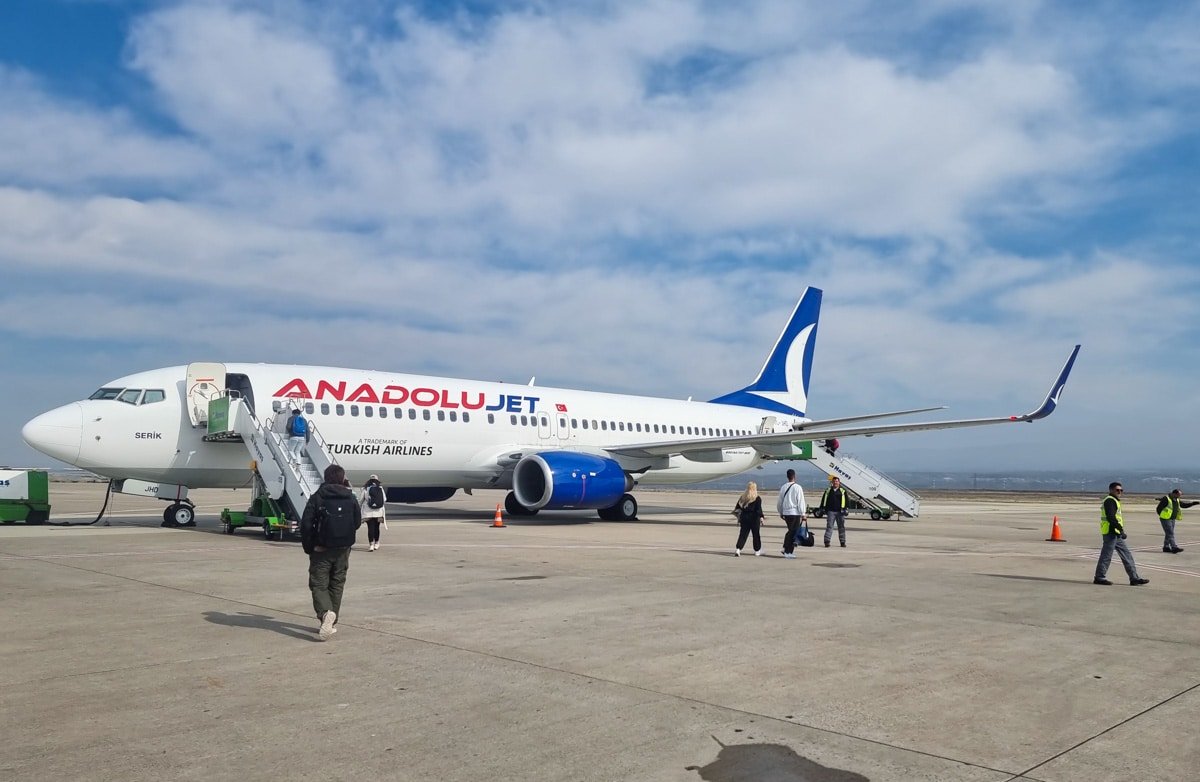A Turkey transport Anadolujet airplane parked on a tarmac with ground crew workers and passengers near the boarding stairs under a clear sky.