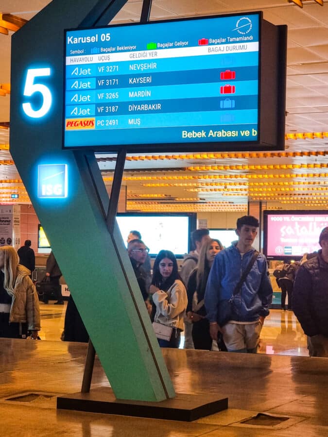 People standing and walking in an airport terminal in Turkey, with a digital flight information display board in the background.