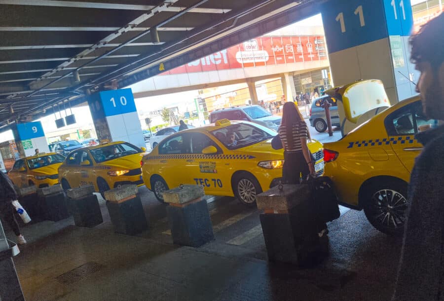 Yellow taxis lined up at a busy airport terminal in Turkey with passengers loading luggage, numbers 9 to 11 visible overhead.