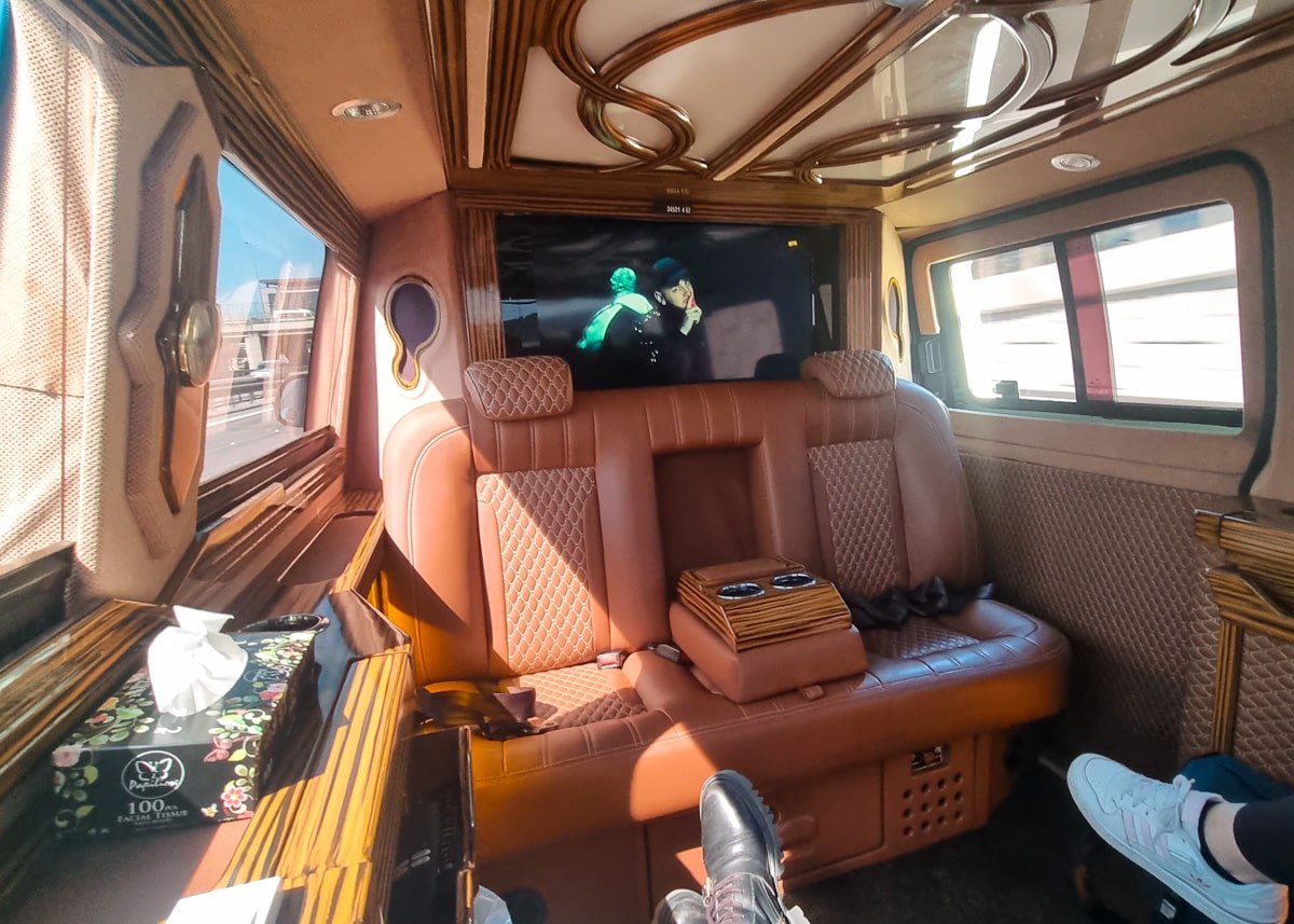 Interior of a luxury transport van with brown leather seats, dark wood accents, and a mounted tv screen displaying a movie.