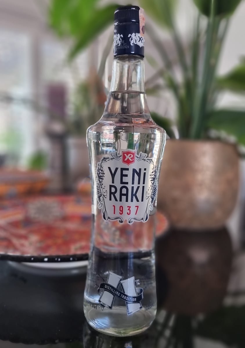 A clear bottle of Yeni Raki, a Turkish spirit, set on a table with plants softly blurred in the background. The label prominently displays "1937.