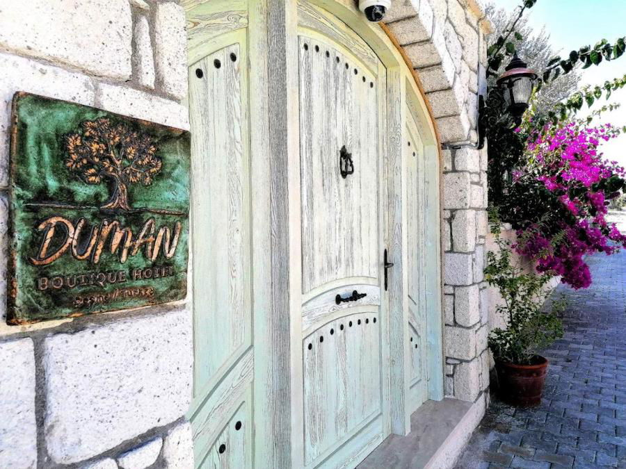 Entrance to Çeşme duhan boutique hotel with a weathered wooden door, flanked by stone walls and flowering plants.