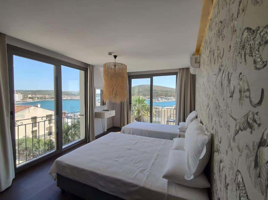 A bright hotel room in Çeşme with a large bed, animal-print wallpaper, and a view of the sea through sliding glass doors.