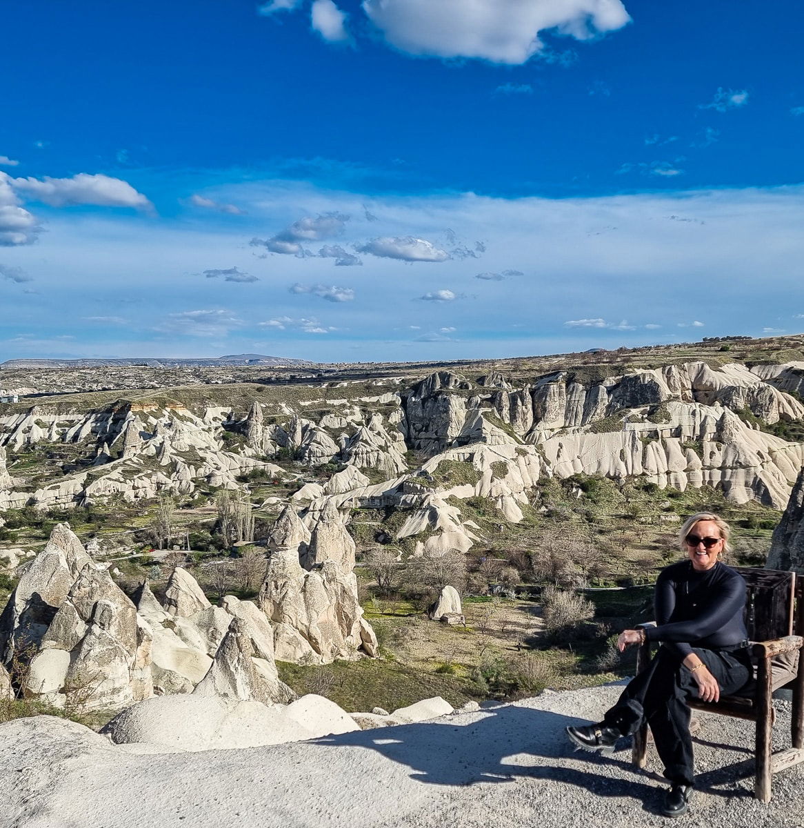 A woman sitting on a wooden chair, smiling at the camera, with the rocky landscape of Goreme, Cappadocia, in the background.