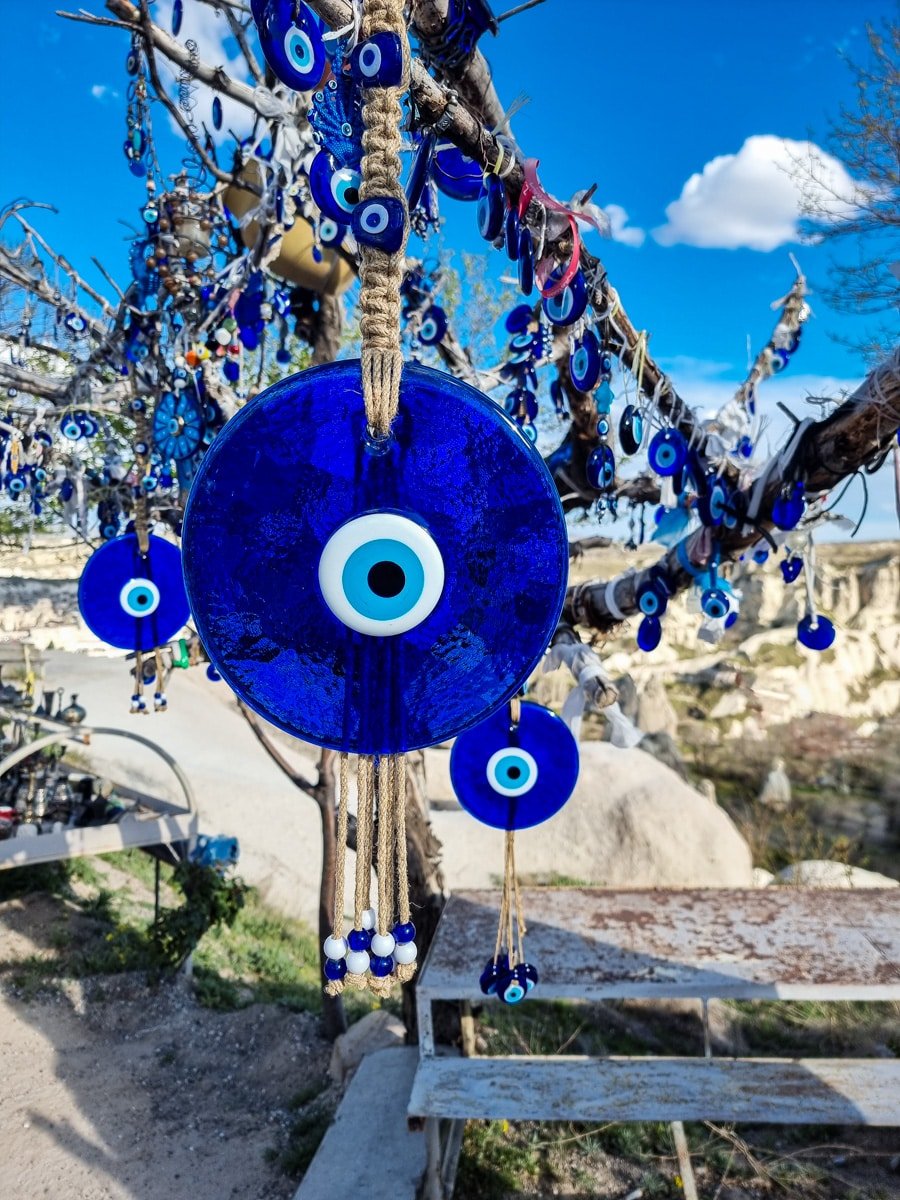 Large blue evil eye amulet hanging on a tree with multiple similar charms in Goreme, set against a clear sky and rocky landscape.