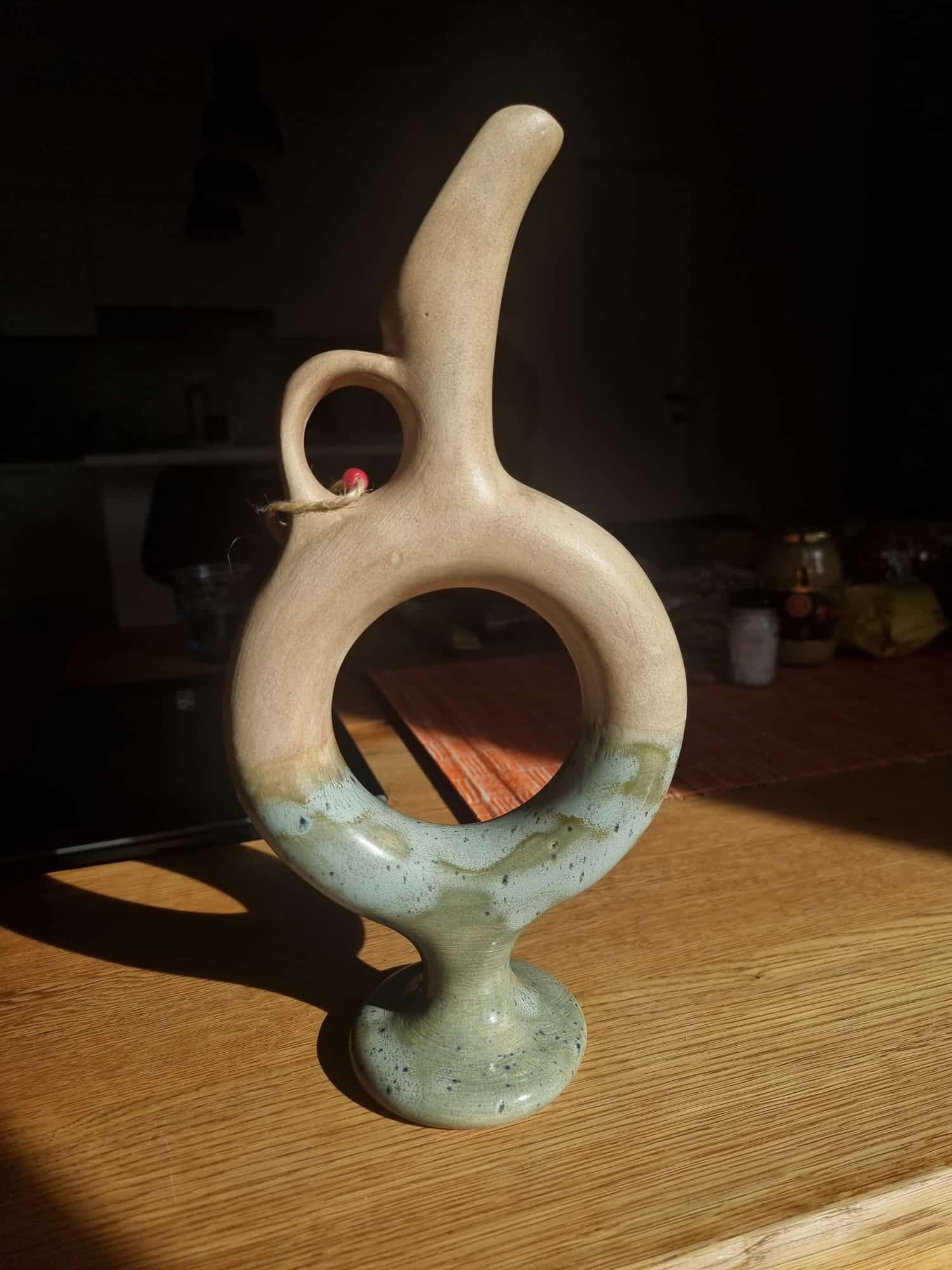 A ceramic pitcher with a unique loop handle and a speckled blue base stands on a wooden table, bathed in sunlight.