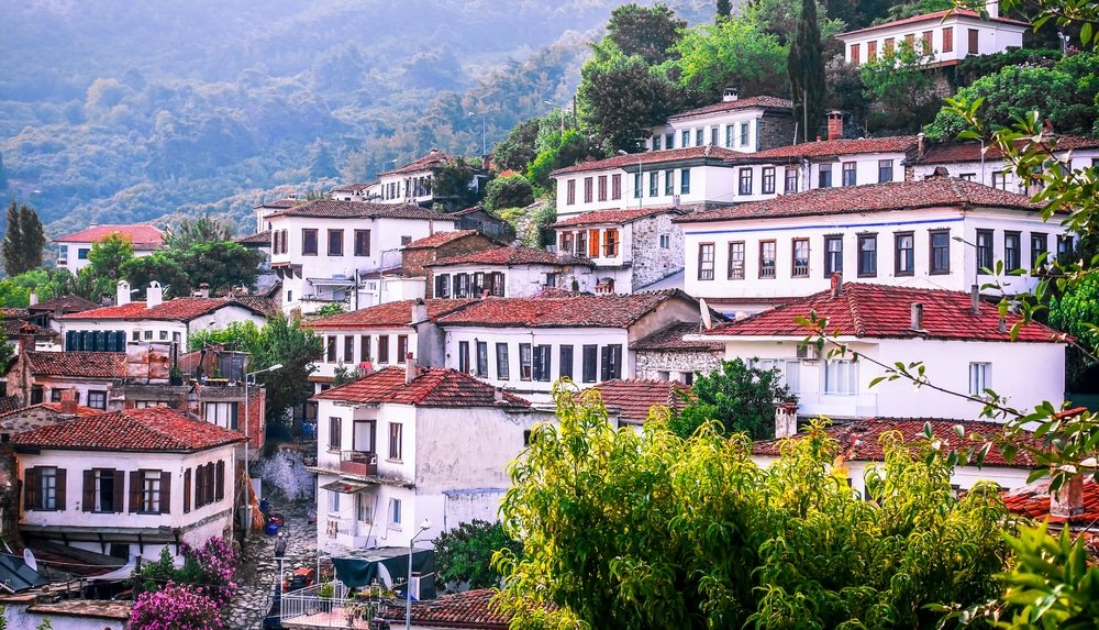 Lush greenery surrounds traditional white houses with tiled roofs in a hilly village in Aydın Province.