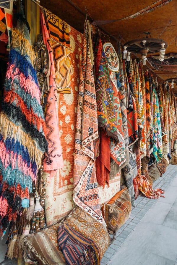 Colorful, patterned rugs and fabrics hanging on display at an outdoor market stall along a sidewalk showcase just one delightful way to immerse yourself in local culture. For those wondering how to spend 3 days in Cappadocia, exploring such vibrant markets is a feast for the senses.