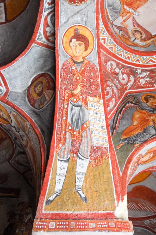 A fresco painting of a saint in red and white robes holding a scroll, adorned with a halo. The artwork is on a pillar, with additional religious scenes visible on the surrounding ceiling—a must-see for anyone figuring out how to spend 3 days in Cappadocia.