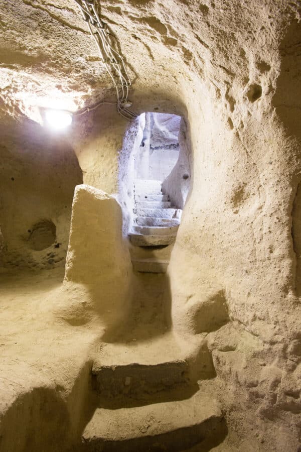 View of an underground tunnel with stone steps leading up to another level, illuminated by a light bulb. The tunnel walls are rough and the ceiling has visible wiring. It's reminiscent of exploring hidden passages when figuring out how to spend 3 days in Cappadocia.