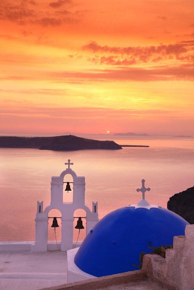 A blue-domed building with bell towers is set against the backdrop of Firostefani and the tranquil sea and an orange-hued sunset, making it one of the best photo spots in Santorini.