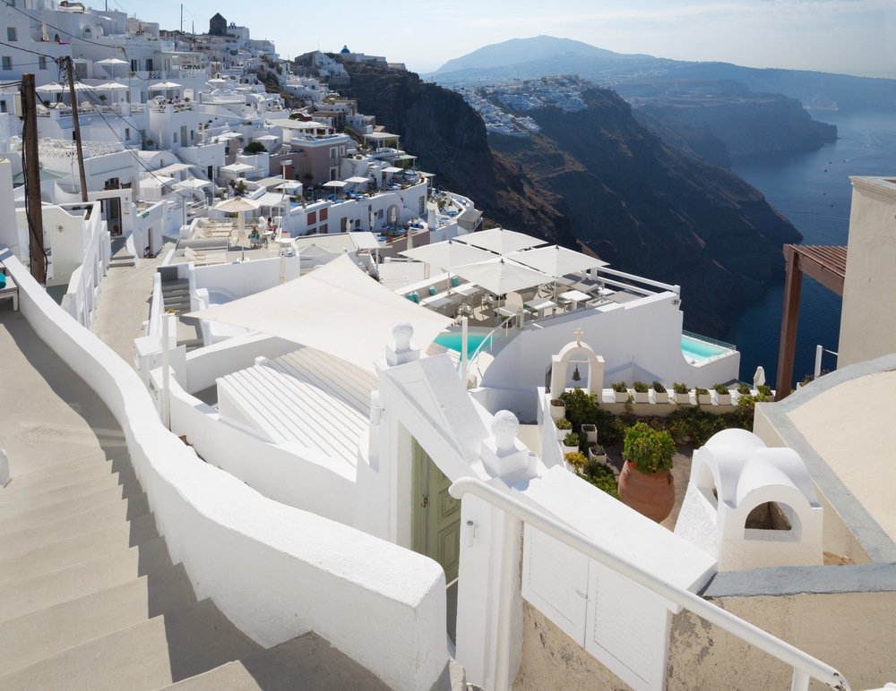 A scenic view of white buildings on a cliffside in Imerovigli, Santorini, Greece, overlooks the Aegean Sea. The landscape includes terraced structures, pathways, and a distant mountainous backdrop. For those seeking the best photo spots, this picturesque setting comes highly recommended.