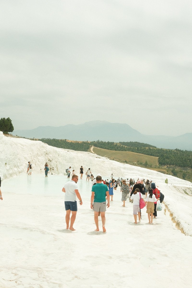 People walk on white travertine terraces filled with water at Pamukkale, Turkey. This breathtaking landscape, which includes distant mountains under a cloudy sky, is truly worth visiting.