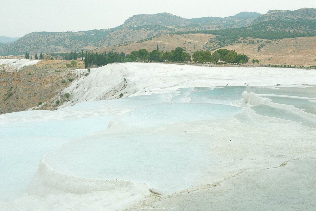 Terraced pools of water surrounded by white travertine formations at Pamukkale, set against a backdrop of hills and sparse vegetation, make it a place worth visiting.