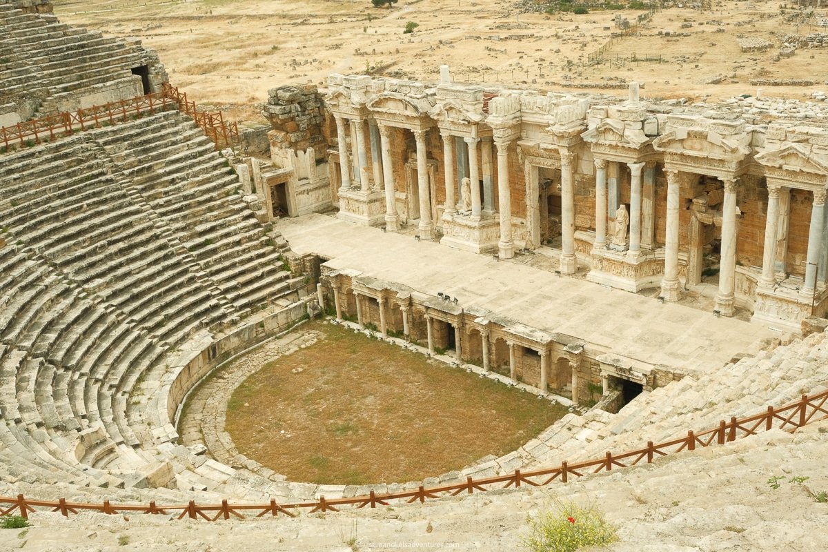 An ancient amphitheater with tiered stone seating and a partially preserved stage area, surrounded by a dry, grassy landscape, offers a glimpse into history when visiting Pamukkale & Hierapolis