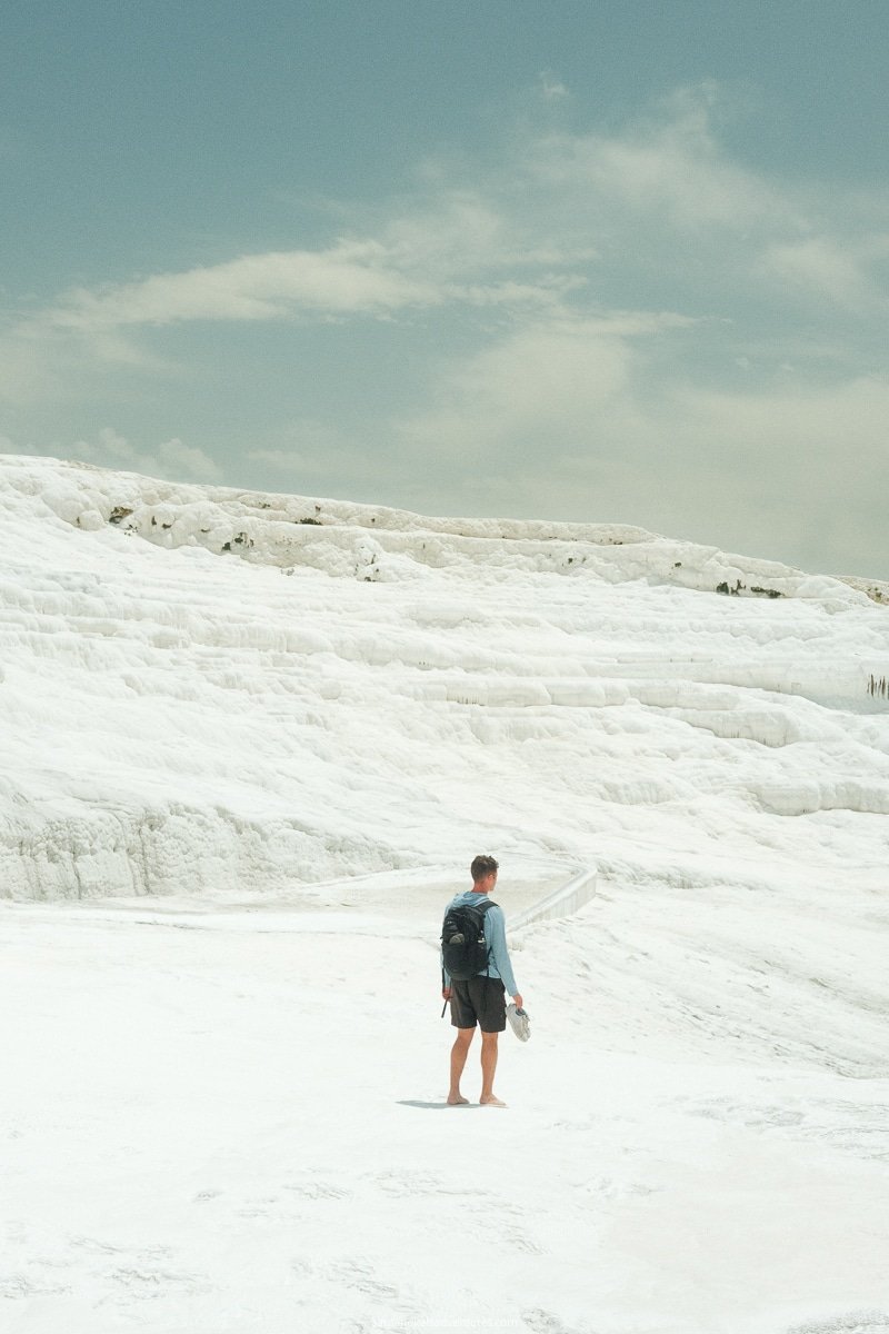 A person in shorts and a long-sleeve shirt, carrying shoes and a backpack, stands on the vast white travertine terrace of Pamukkale under a partly cloudy sky—truly worth visiting.