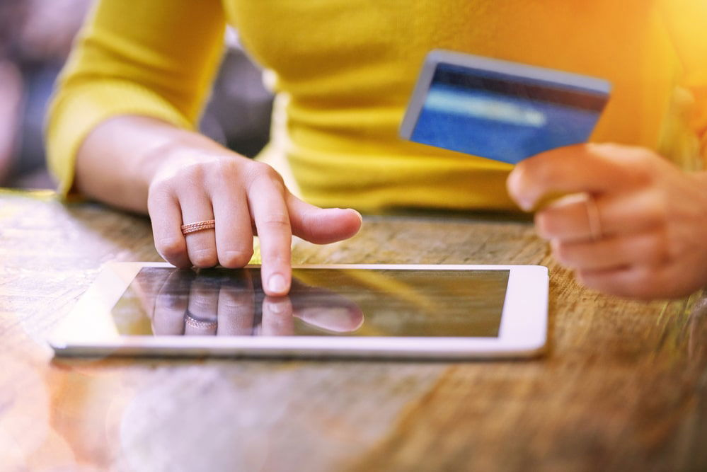 A person in a yellow sweater uses a tablet while holding a credit card, managing travel expenses during peak season.