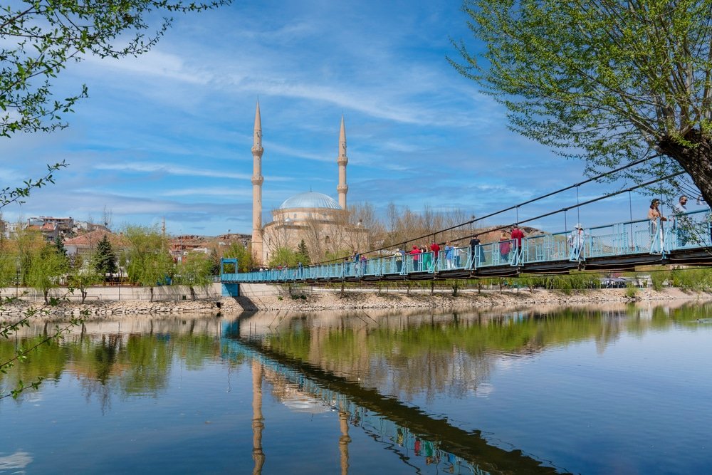 Strolling across the suspension bridge over the Kızılırmak River, with the idyllic backdrop of Avanos in Cappadocia. A stunning mosque with two minarets stands tall, framed by tree branches, in this picturesque pottery village.