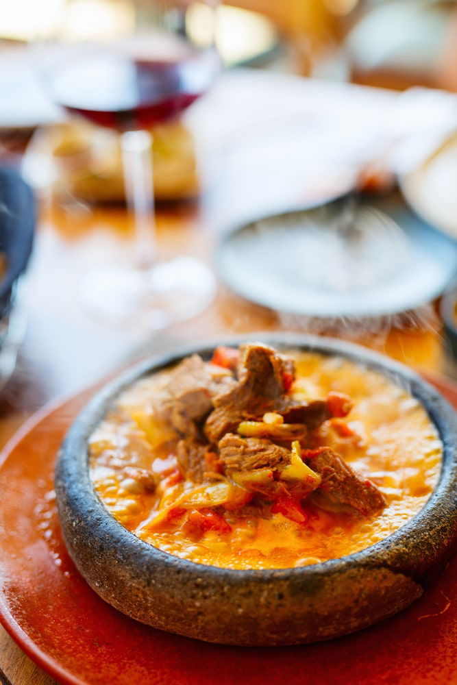 A close-up of a bowl of stew with chunks of meat and vegetable pieces in a simmering broth, reminiscent of the hearty dishes you might savor when exploring how to spend 3 days in Cappadocia. A glass of red wine is visible in the blurred background.