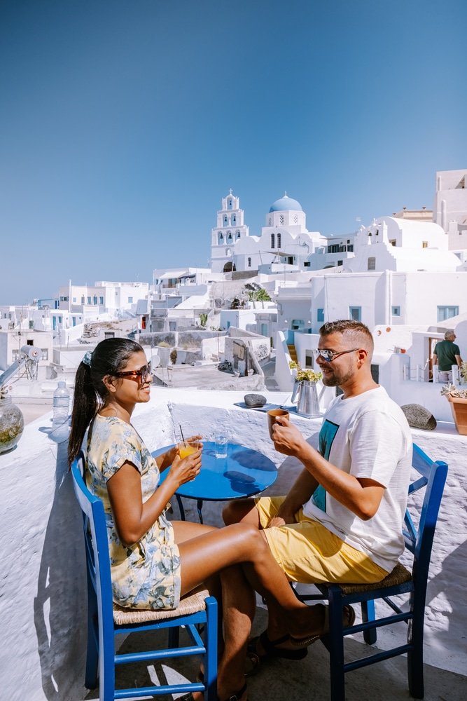 Us sitting in at a table enjoying drinks with a picturesque view of white buildings and blue domes in Pyrgos Santorini, Greece, one of the best photo spots in Santorini.