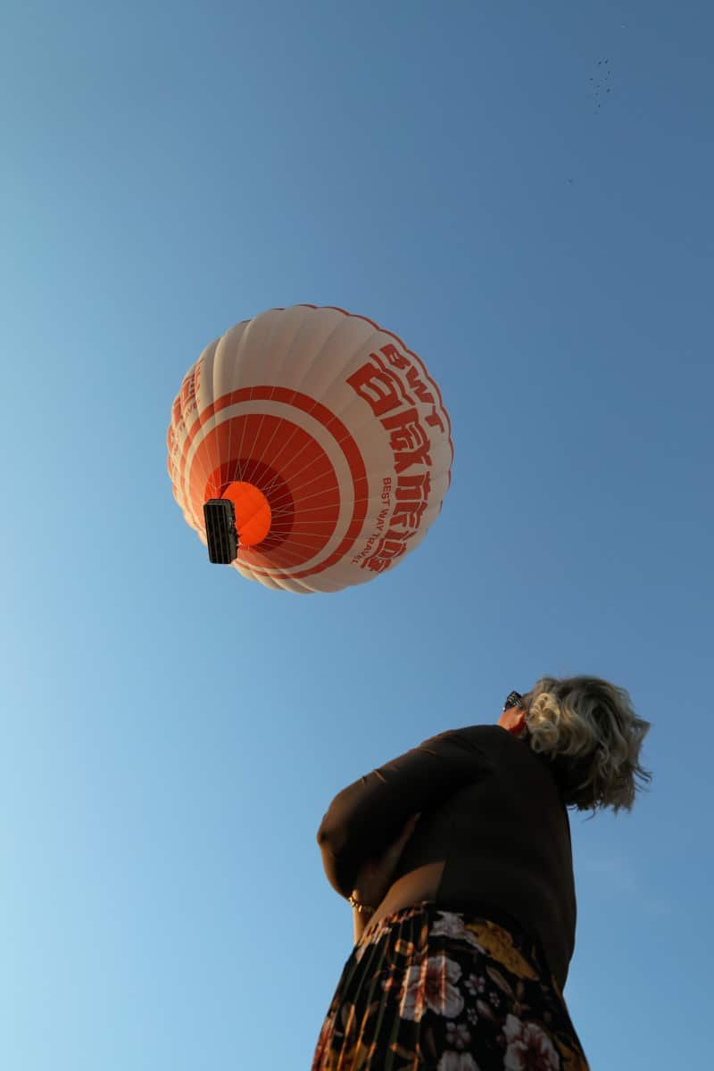 SJ looks up at a large orange and white hot air balloon flying in a clear blue sky, capturing the essence of "How To Spend 3 Days In Cappadocia