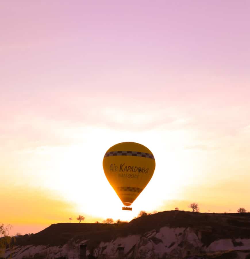 A hot air balloon with "How To Spend 3 Days In Cappadocia" written on it floats in a pastel-colored sky during sunrise.