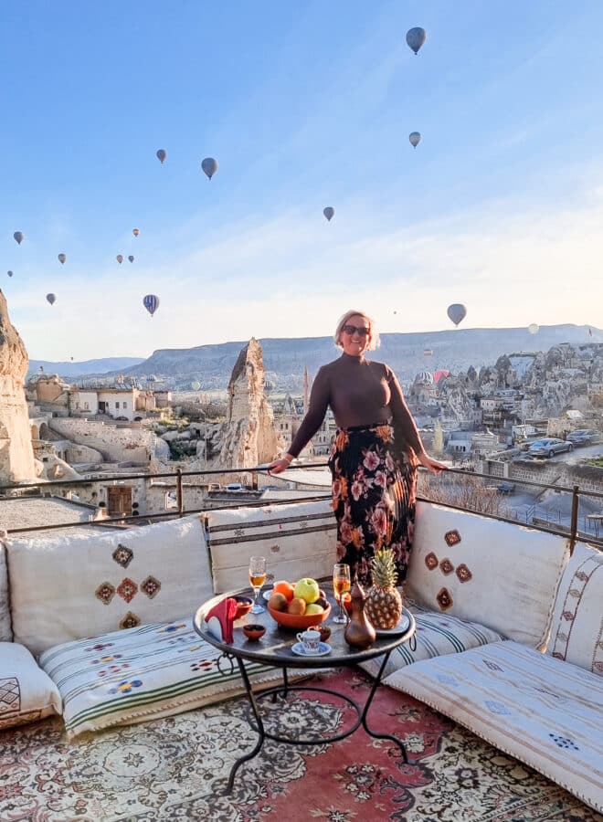 A woman standing on a terrace with a fruit platter, overlooking how to spend 3 days in Cappadocia with balloons rising above rocky formations at sunrise.