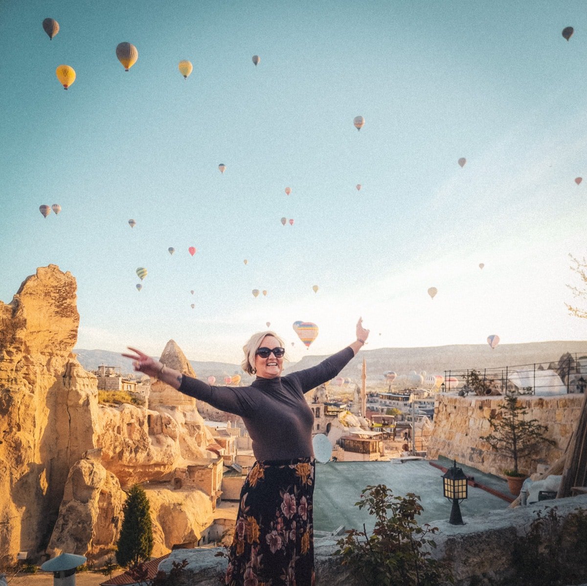 SJ with outstretched arms smiling at the camera, surrounded by colorful hot air balloons above rocky terrain under a clear sky in Cappadocia.