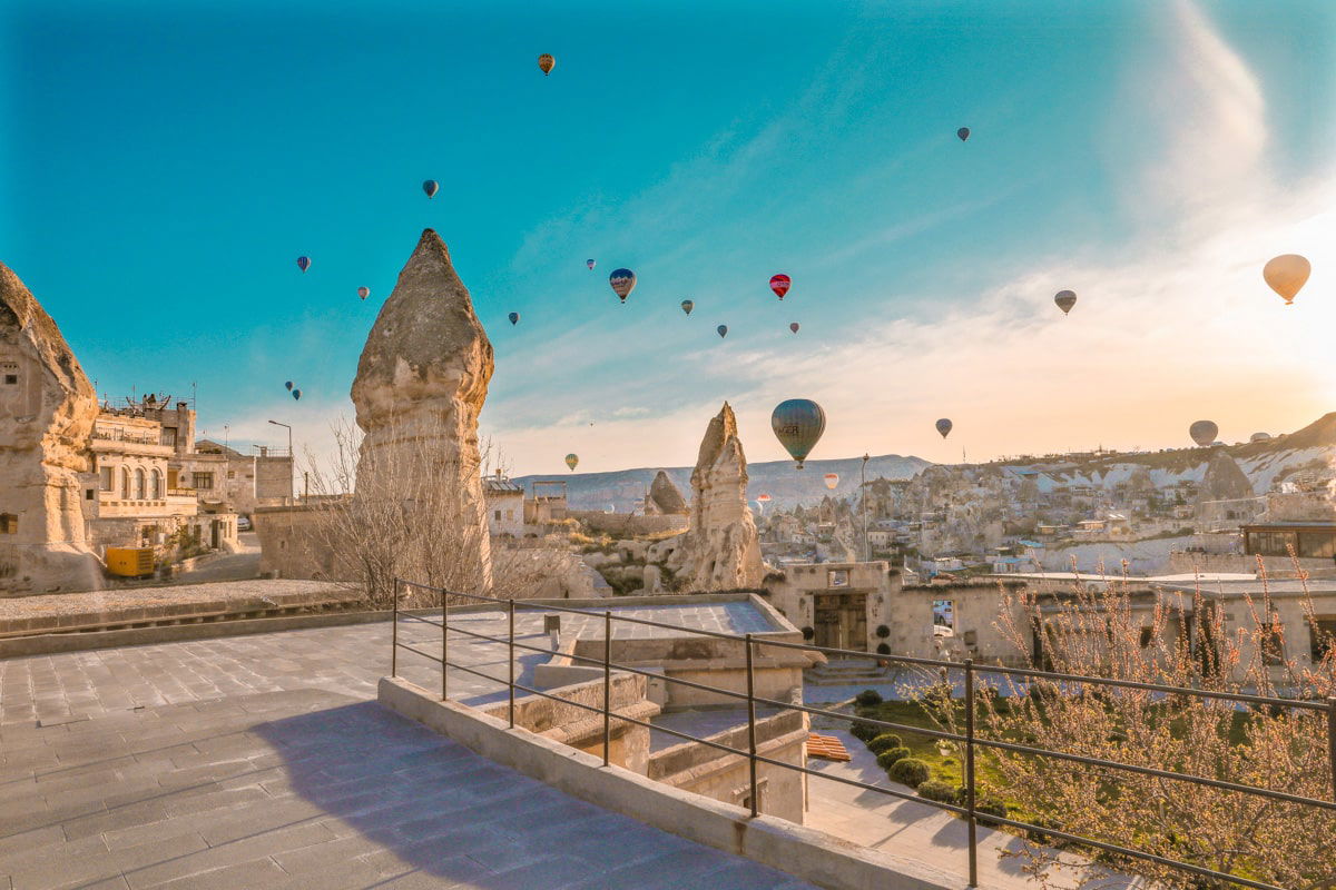 Hot air balloons float above a rocky landscape with unique geological formations under a clear, sunlit sky, showing you how to spend 3 days in Cappadocia, Turkey.