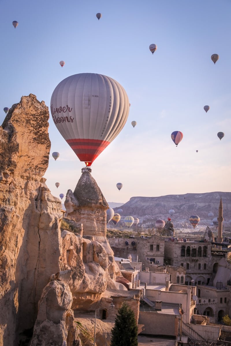 Hot air balloons floating over a scenic rocky landscape with unique rock formations and buildings in Cappadocia at sunrise.