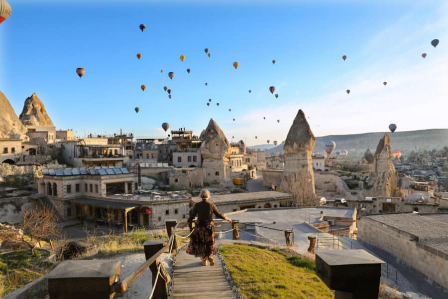 A woman walks down steps in Cappadocia, Turkey, with hot air balloons flying over rock formations and houses at sunrise, capturing the essence of how to spend 3 days in Capp