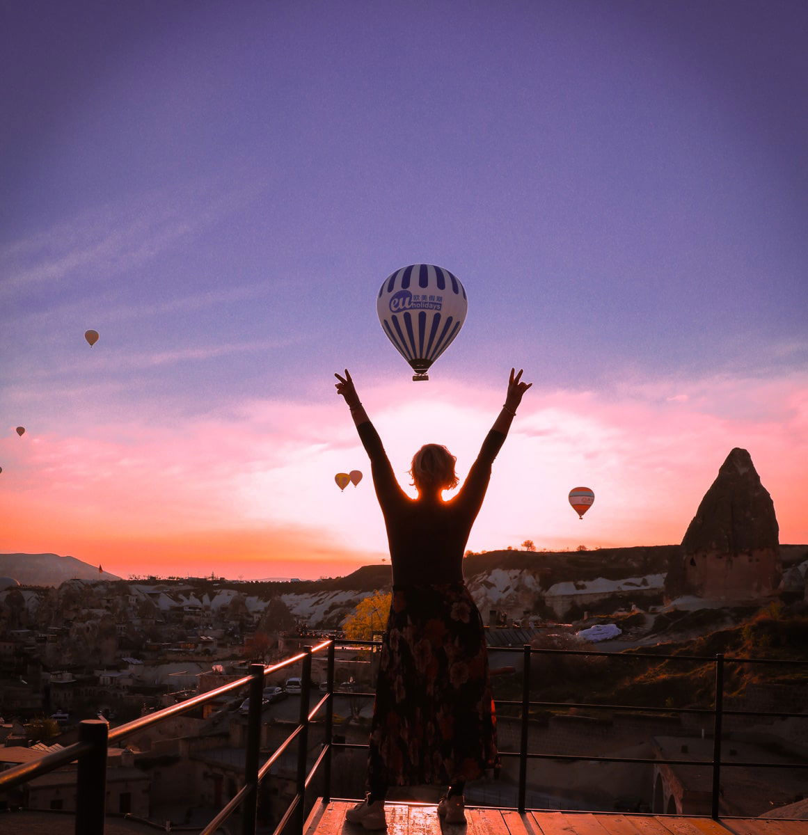 SJ raises her arms towards a sky dotted with hot air balloons at sunset, standing on a balcony overlooking the rocky landscape of Cappadocia.