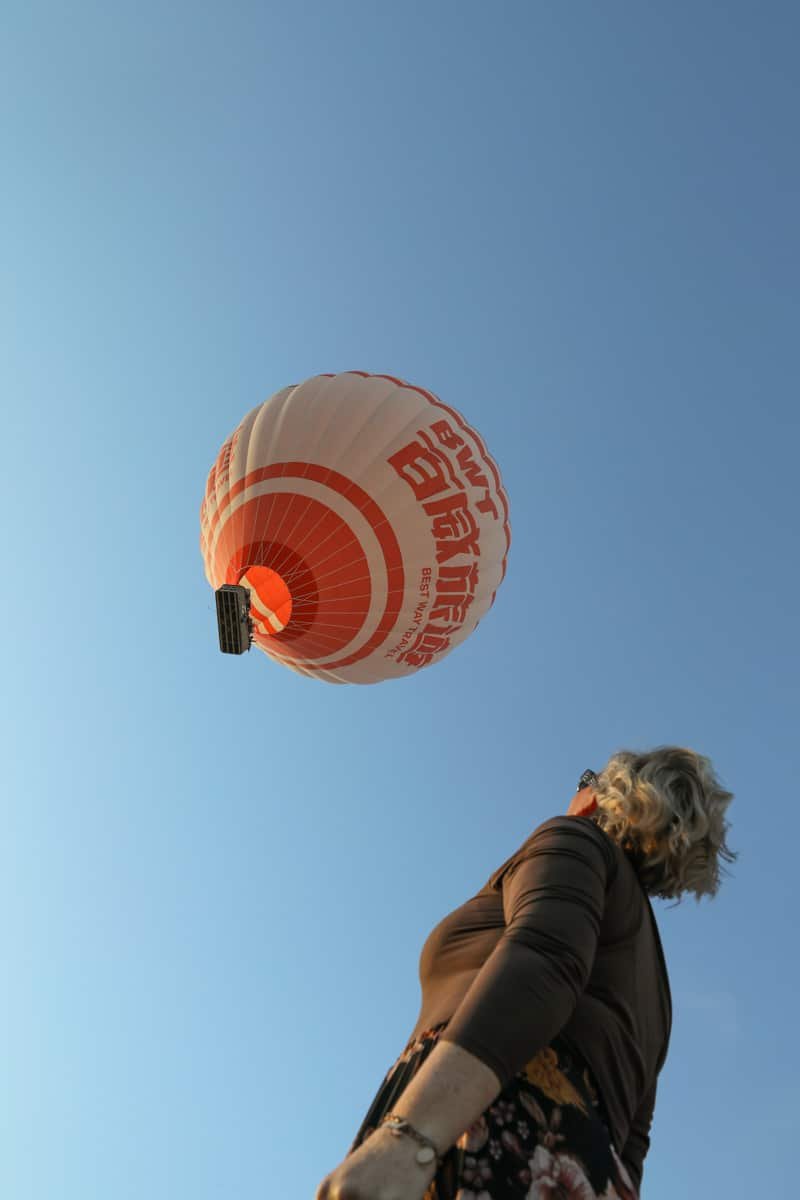 SJ looks up at a large red and white hot air balloon flying against a clear blue sky in Cappadocia.