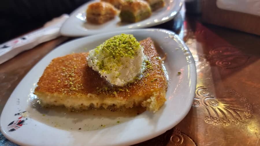 Close-up of a piece of Kunefe topped with clotted cream and ground pistachios on a white plate, with a second plate of baklava pieces visible in the background on a copper-colored table. A taste straight out of "A Guide To Kadikoy Istanbul.