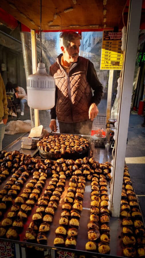 A street vendor in Kadikoy, Istanbul stands behind a table filled with roasted chestnuts. He is wearing a quilted vest and is tending to the chestnuts, with a price list displayed above his head.