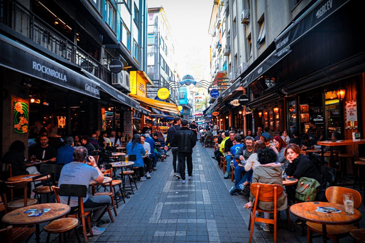 Vibrant street scene with people dining at outdoor cafes, colorful signs, and a man walking down the center in an urban setting.