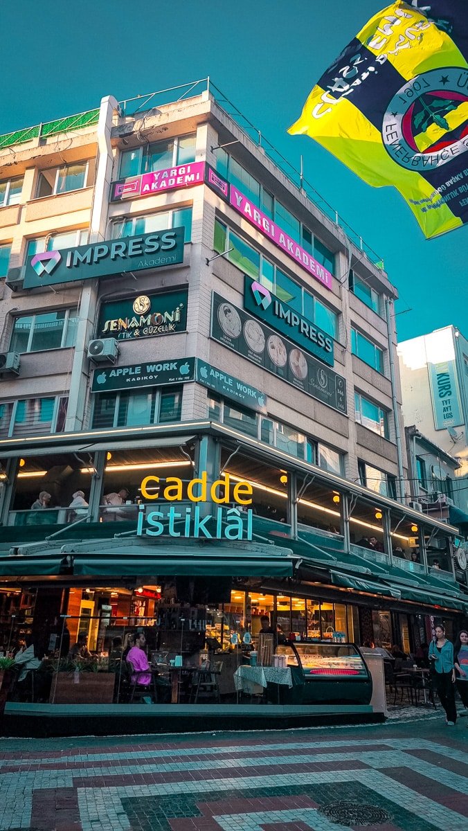 Street view of a multi-story building with shops and banners, including "cadde istiklal" restaurant on the ground floor and several other businesses on the upper floors. Pedestrians wander below a flag above, capturing the essence of A Guide To Kadikoy Istanbul.