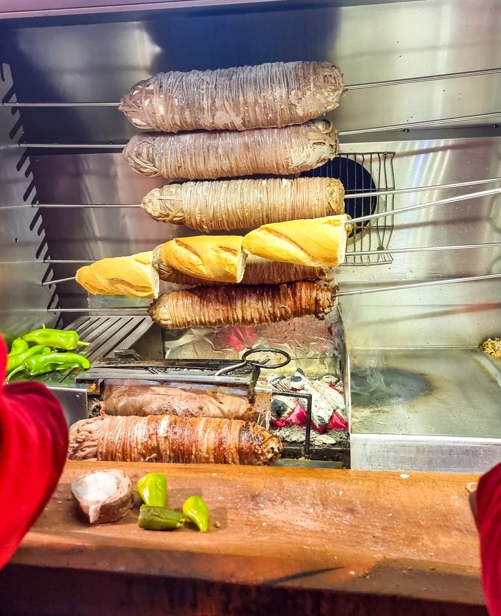 Six large meat rolls cooking on a vertical rotisserie with bread loaves and some bell peppers placed nearby, all inspired by A Guide To Kadikoy Istanbul. An open flame and grill can be seen below the meat.