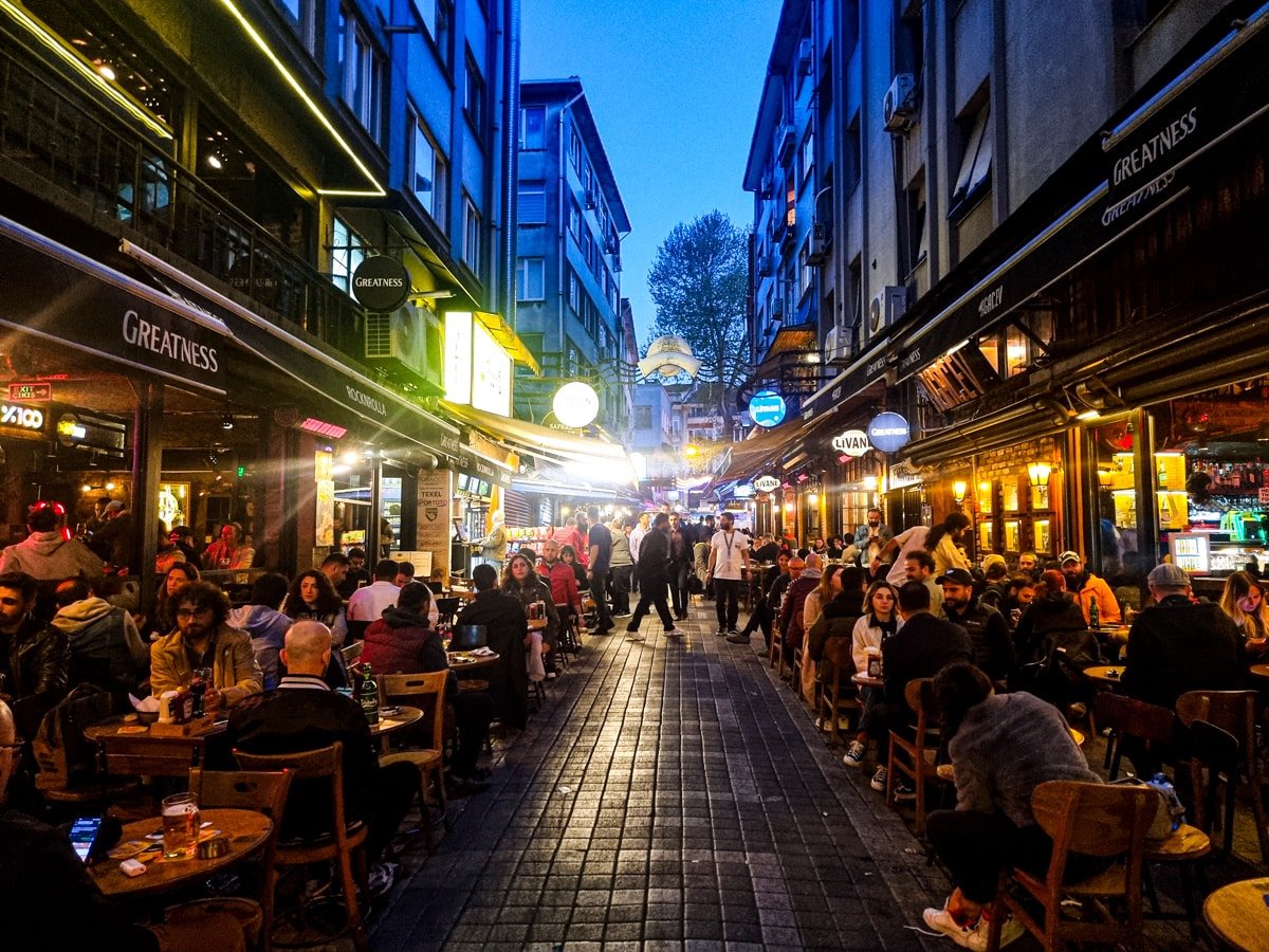 A bustling narrow street at dusk, lined with cafes and restaurants, filled with people dining and socializing. The vibrant scene is illuminated by streetlights and storefront signs—a perfect introduction for anyone exploring A Guide To Kadikoy Istanbul.
