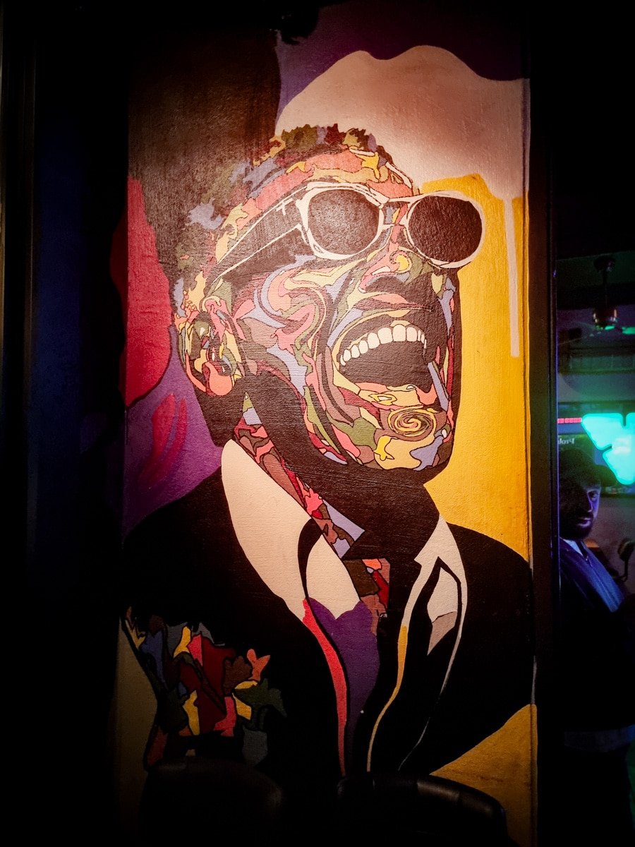 A colorful mural depicting a person wearing sunglasses and laughing, with vibrant patterns on the face and suit, set against a yellow and purple background, bringing to mind the vibrant energy of Kadikoy in Istanbul.